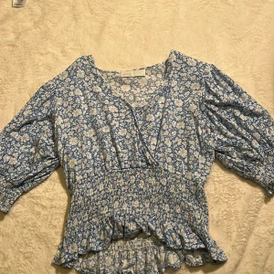 Blommig blus från Samsa Collection - Supersöt blommig blus i blått och vitt från Samsa Collection. Blusen har v-ringning, smockad midja och volangkant nertill. Trekvartsärmar med resår vid ärmslut ger en puffig känsla. Perfekt för dig som gillar romantisk och feminin stil.