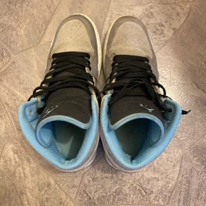 Nike Air Jordan 1 Mid grå/blå sneakers - Nike Air Jordan 1 Mid sneakers i grått tyg med svarta detaljer och svart snörning. Insidan är ljusblå och swoosh-loggan har ett glittrigt mönster. Klassisk hög modell med rund tå och perforerad tåbox. Perfekta för dig som gillar streetstyle. Ingen låda och inget kvitto men äkthet garanteras alltid hos oss! 