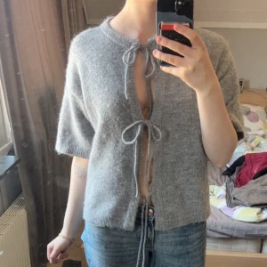 Grå stickad kofta med knytband - Säljer en grå stickad kofta med kort ärm och två knytband framtill. Koftan har en mjuk och fluffig känsla och är perfekt att slänga över ett linne eller t-shirt. Enkel och stilren design som passar till mycket.