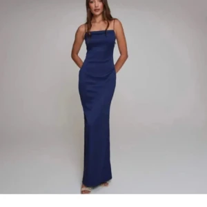 Marinblå långklänning med smala axelband - Elegant marinblå långklänning med smala axelband och figurnära passform. Klänningen har rak halsringning och öppen rygg, vilket ger en stilren och modern look. Perfekt för speciella tillfällen.