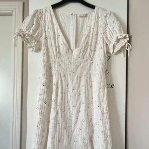 Vit blommig kortklänning från Kiss The Sky - Supersöt vit kortklänning från Kiss The Sky med små gula blommor. Använd en kväll!