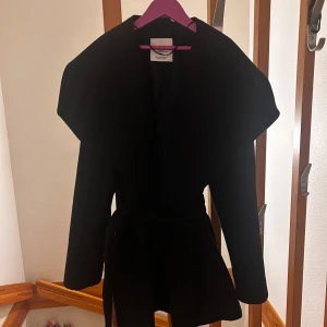 Kappa från mango  - Elegant svart kappa från MNG Suit Collection, storlek XS (EUR S). Stilren design med bred krage och knytskärp i midjan – perfekt för både vardag och finare tillfällen. Knappt använd och i mycket fint skick. Nypris ca 1200 kr. Öppen för rimliga prisförslag – först till kvarn!