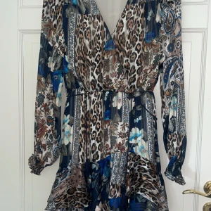 Klänning från  Guess - Säljer en snygg långärmad klänning från Guess med blandat leopard- och blommönster i blått, beige och brunt. Klänningen har v-ringning, volangdetaljer och markerad midja. Perfekt för dig som vill sticka ut med en unik stil. 💕 endast använd en gång! 