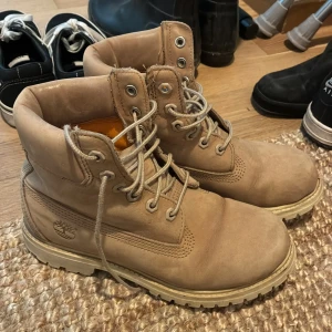 Beige boots från Timberland - Säljer ett par klassiska beige boots från Timberland med grov sula och snörning. Skorna har rund tå och är tillverkade i skinn, perfekta för höst och vinter. Ikonisk Timberland-logga på sidan.