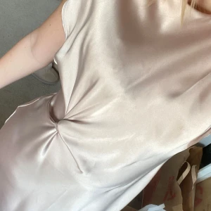 Champagnefärgad satinklänning med twistdetalj - Säljer en elegant långklänning i champagnefärgad satin med korta ärmar och snygg twistdetalj i midjan. Klänningen har en mjuk, glansig yta och en liten öppning i ryggen vid knappen. Perfekt som balklänning eller till finare middagar! Går nästan hela vägen ner på mig som är 173. Den har även en väldigt liten fläck lite längre ner på ryggen, men det tänker man inte på.
