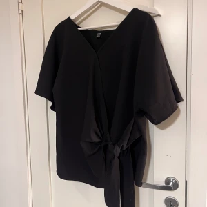 Svart blus med knytband - Säljer en svart blus med v-ringning och korta ärmar. Blusen har ett brett knytband i midjan som ger en snygg detalj och markerar midjan. Xl men sitter som en L/M