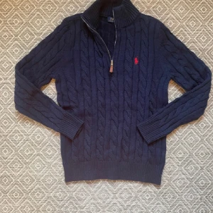 Mörkblå stickad tröja med dragkedja från Polo Ralph Lauren - Mörkblå kabelstickad tröja från Polo Ralph Lauren med halv dragkedja och röd broderad logga på bröstet. Hög krage och långa ärmar. Använd cirka 2 månader. 