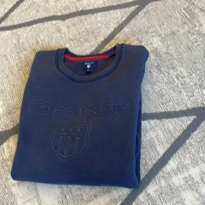 Mörkblå sweatshirt från GANT - Säljer en mörkblå sweatshirt från GANT med broderad logotyp och sköld på bröstet. Tröjan har rund halsringning och långa ärmar. Klassisk och stilren modell med ribbade muddar vid ärmslut och nederkant.