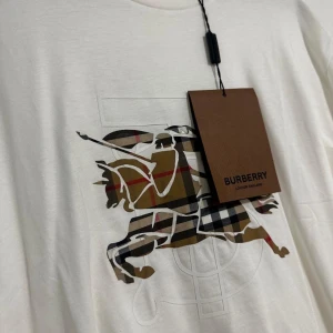 Vit t-shirt med Burberry-ryttare - Vit t-shirt från Burberry med klassiskt ryttarmotiv i märkets rutiga mönster på bröstet. Rund halsringning och korta ärmar. Perfekt för dig som gillar stilrena plagg med lyxig känsla.