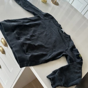 Svart sweatshirt från Stone Island - Säljer en svart sweatshirt från Stone Island junior med klassisk logotyp-patch på ärmen. Tröjan har rund halsringning och långa ärmar. Perfekt för en stilren och avslappnad look.