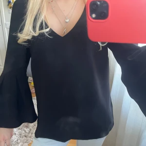 Svart blus med vida armar - Snygg svart blus med v-ringad hals och vida trumpetärmar. Perfekt att styla med jeans eller kjol för en trendig look. Tunt och mjukt material som faller fint.