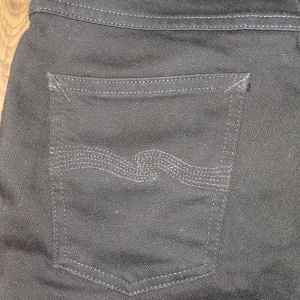 Svarta grim tim Nudie Jeans  - Säljer ett par svarta jeansbyxor från Nudie Jeans med klassisk femficksdesign och diskret broderad bakficka. Byxorna har normal passform och raka ben, perfekta till vardags. Stängs med dragkedja och knapp. De är storlek W30 L32 dem är inte andvänd så mycket jag är prutbar vid snabbaffär skriv om du har några frågor eller funderingar!