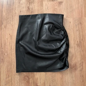 Snygg svart kjol i skinnimitation från Zara - Säljer en svart kortkjol från Zara i skinnimitation med snygg rynkdetalj på ena sidan. Kjolen har en tight passform och är tillverkad i polyester. Perfekt för dig som gillar en edgy och trendig stil.