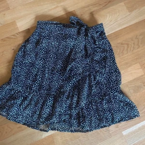 Svart omlottkjol med vita prickar - Säljer en svart omlottkjol med vita prickar och volangkant. Kjolen har en knytning i midjan och är kort i modellen. Perfekt för dig som gillar en söt och trendig stil.