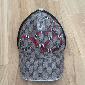 Gucci keps snake  - Snygg keps från Gucci med klassiskt GG-mönster i grått och svart samt en röd, vit och svart orm på framsidan. Baksidan är i svart mesh och justerbar med ett band i grönt och rött. Perfekt för att sticka ut i mängden. Pris kan diskuteras vid snabb affär.