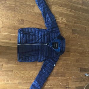Blå dunjacka från Sail Racing - Säljer en blå dunjacka från Sail Racing med vit dragkedja och hög krage. Jackan har quiltad design och ett Sail Racing-märke på ärmen. Perfekt för kyligare dagar och har en sportig look.