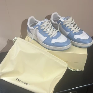 Blå och vita sneakers från Axel Arigato - Säljer ett par fräscha sneakers från Axel Arigato i blått och vitt. Skorna har klassisk design med snörning och platt sula. Perfekta för dig som vill ha en stilren och modern look. Skorna kommer med originalpåse.