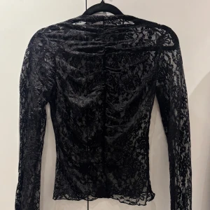 Svart spetsblus från Gina Tricot - Säljer en svart långärmad spetsblus från Gina Tricot. Blusen är genombruten med blommigt spetsmönster och har en tight passform. Perfekt att styla lager på lager eller med ett linne under.