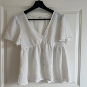 Vit blus med v-ringning och kort ärm - Säljer en söt vit blus med v-ringning och korta ärmar. Blusen har ett diskret broderat prickmönster och lätt volang nedtill. Perfekt till sommaren och passar till både jeans och kjol. Använt 1 gång.