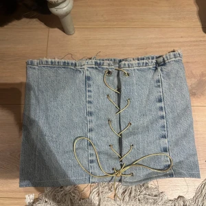 Jeans tubtopp med snörning - Säljer en snygg tubtopp i ljusblå denim med snörningsdetalj framtill. Toppen är axelbandslös och har en cool, croppad passform. Perfekt att styla till både kjol och byxor för en trendig look.
