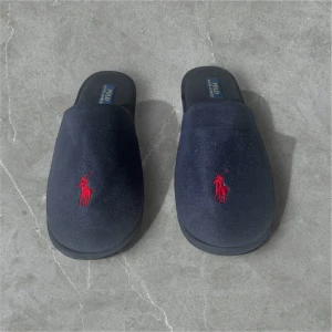Mörkblå tofflor från Polo Ralph Lauren - Mörkblå tofflor från Polo Ralph Lauren med röd broderad logga framtill. Mjukt material och platt sula för extra komfort. Perfekta att glida runt i hemma. Storlek 46
