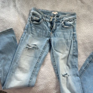 Ljusblå slitna jeans med bootcut - Säljer ett par ljusblå jeans med bootcut och slitna detaljer på benen. Jeansen har klassisk femficksdesign, dragkedja och knapp framtill. Perfekta för en avslappnad och trendig look.