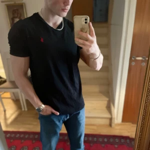 Svart t-shirt från Polo Ralph Lauren - Svart t-shirt från Polo Ralph Lauren med klassisk passform och liten röd broderad logga på bröstet. Perfekt basic-plagg med v-ringad hals och korta ärmar. Tillverkad i mjuk bomull.