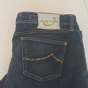 Mörkblå jeans från Jacob Cohen - Snygga mörkblå jeans från Jacob Cohen med klassisk femficksdesign och kontrastsömmar. Byxorna har en vit patch med guldlogga bak i midjan och gylf med knapp. Perfekta för dig som gillar stilrena och exklusiva jeans.
