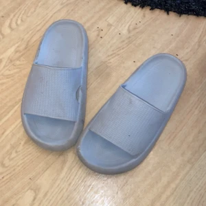 Gråa sandaler i syntetmaterial - Ett par enkla gråa sandaler i syntetmaterial med bred rem över foten och platt sula. Perfekta att glida runt i hemma eller på stranden. Lätta att ta på och av.