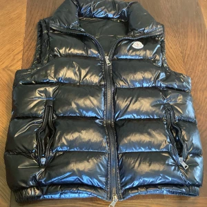 Svart dunväst från Moncler - (ALLT FUNKAR) Säljer en svart, glansig dunväst från Moncler med hög krage och dragkedja framtill. Västen har två fickor med dragkedja och klassisk Moncler-logga på bröstet. Perfekt för lager-på-lager under kyliga dagar.