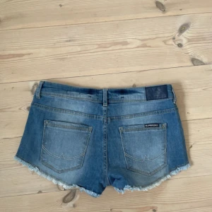 Blå jeansshorts med fransar och nitar - Säljer ett par blå jeansshorts från Superdry med fransig kant och snygga nitar längs fickorna. Shortsen har normal passform, klassiska fickor och låg midja. Perfekta för sommaren och ger en cool, avslappnad look. Skriv för fler bilder, och kolla gärna in de andra shortsen jag säljer! 