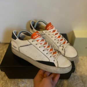 Vita sneakers från Crime London med orange detaljer - Säljer ett par vita sneakers från Crime London med svarta och orange detaljer. Skorna har snörning med vita skosnören, rund tå och platt sula. Märkeslogga på plösen och hälen. Perfekta för en cool och avslappnad stil. Storleken är 40,5 men passar för 41. Dom är ca 6/10 i skick pga en spricka i sulan som syns på bild 4. Fråga på om ni har frågor.🙂