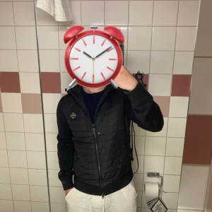 Svart quiltad hoodiejacka med dragkedja - Säljer en svart cardigen från J.Lindberg med quiltad framsida och rygg, samt stickade ärmar. Jackan har dragkedja, huva och två sidofickor med dragkedja. Perfekt för dig som gillar en sportig och stilren look.