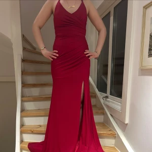 Röd långklänning med öppen rygg och slits - Säljer en elegant röd långklänning med smala axelband och djup v-ringning. Klänningen har en hög slits framtill och en öppen rygg med snygg snörning. Perfekt för bal eller fest. Materialet är mjukt och följsamt. Använd en gång. På sista bilden kan man se att den har omärkbara fläckar men dom syns knappt! 