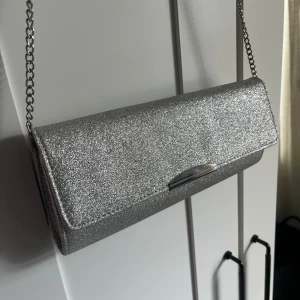 Glittrig silver kuvertväska med kedja från Anna Field - Säljer en snygg kuvertväska i glittrigt silver med kedja från Anna Field. Väskan har ett stilrent metallspänne och är perfekt för att lyfta din outfit. Insidan har ett litet fack för småsaker. Superfin att bära crossbody eller i handen. Funkar alltså även utan kedja som man kan lägga i väskan om man vill ha till en bal eller liknande💞Har några lätta missfärgningar, men inget jag tycker man lägger märke till om man inte kollar världigt noga, därför jag säljer den billigt. Skriv för fler bilder🩷