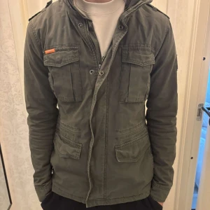 Olivgrön militärinspirerad jacka från Superdry - Säljer en olivgrön Superdry Rookie Edition jacka med fyra fickor framtill, axelklaffar och dragkedja med knappslå. Jackan har en klassisk militärlook och är tillverkad i bomull. Perfekt för dig som gillar streetwear och funktionella detaljer.