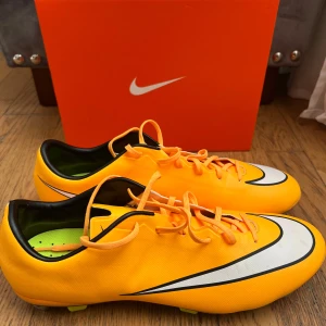 Nike fotbollsskor i orange - Säljer ett par snygga orangea Nike fotbollsskor med svart och vit detalj på sidan. Skorna har snörning och rund tå, samt dobbar för bra grepp på planen. Perfekta för dig som vill sticka ut på fotbollsplanen!