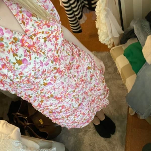 Blommig kortklänning med volang - Säljer en söt kortklänning med blommigt mönster i rosa, orange och grönt på vit botten. Klänningen har v-ringning, korta ärmar och volangdetaljer längs kjolen. Perfekt för sommarens tillfällen!