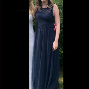 Mörkblå långklänning med spetsdetaljer - Säljer en elegant mörkblå långklänning med spets upptill och rund halsringning. Klänningen har ärmlös design och ett flytande, lätt tyg i kjolen som ger en fin siluett. Perfekt för speciella tillfällen som till exempel balen i sommar !!