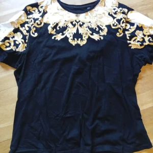 Svart t-shirt med guldmönster - Snygg svart t-shirt med unikt guldmönster upptill och på axlarna. Rund halsringning och korta ärmar. Perfekt för dig som vill sticka ut med en cool och trendig stil.