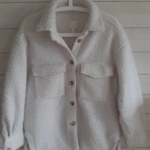 Vit teddyjacka med knappar - Mysig vit teddyjacka med krage och knäppning framtill. Jackan har två bröstfickor med lock och långa ärmar. 
