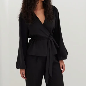 Svart omlottblus från NA-KD - Säljer en elegant svart omlottblus från NA-KD med knytband i midjan. Blusen har långa ärmar och en v-ringad design som ger en stilren look. Perfekt för att klä upp eller ner beroende på tillfälle.