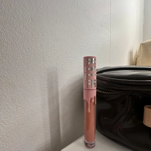 Kylie Cosmetics läppglans i nude - Pris kan diskuteras! Säljer ett läppglans från Kylie Cosmetics i en nude-rosa nyans. Förpackningen har ett rosa lock med droppdesign och stor silvrig logga. Perfekt för att ge läpparna glans och en naturlig färg.