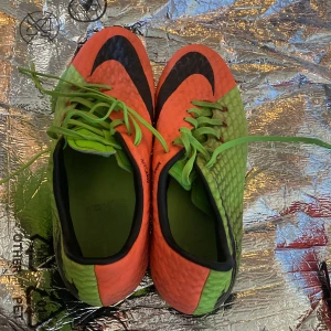 Nike fotbollsskor i orange och grönt - Säljer ett par Nike fotbollsskor med cool färgblockning i orange och grönt. Skorna har snörning och rund tå, samt dobbar för spel på gräs. Perfekta för dig som vill sticka ut på planen!