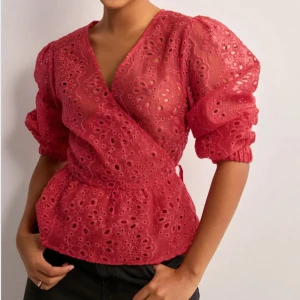 Röd omlottblus med broderade detaljer - Säljer denna röda omlottblus med v-ringning och broderade hålmönster från neo noir. Blusen har puffiga långärmar med resår vid ärmslut och markerad midja. Perfekt för dig som vill ha något unikt och trendigt! Aldrig använd och prislapp finns kvar! 