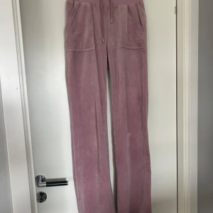 Rosa byxor från Juicy Couture - Rosa byxor från Juicy Couture med raka ben och dragsko i midjan. Byxorna har fickor fram och bak med knappar och klassisk Juicy Couture-logga broderad på ena fickan. Lite fläckar baktill på vaderna (se bild 4).