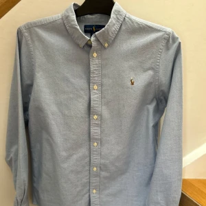 Ljusblå skjorta från Ralph Lauren - Klassisk ljusblå skjorta från Ralph Lauren med broderad logga på bröstet. Skjortan har knappar hela vägen, button-down krage och lång ärm. Perfekt för en stilren och tidlös look. Priset är diskuterbart