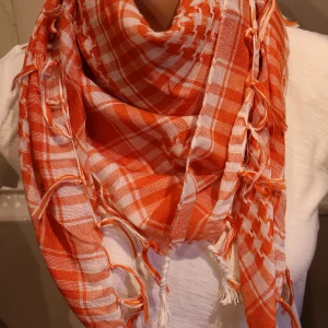 Palestinasjal Orange och vit rutig halsduk med fransar - Säljer en snygg orange och vit rutig halsduk med fransar längs kanterna. Halsduken är mjuk och har ett klassiskt rutmönster som passar perfekt för att liva upp din outfit.
