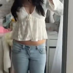 Vit blus med knyt och volang från Ginatricot  - Säljer en söt vit blus med v-ringning, knyt framtill och volang nedtill. Blusen har långa ärmar och är tillverkad i ett lätt, transparent material med blommigt mönster. Perfekt för en fräsch och romantisk look. 