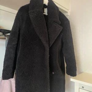 Svart teddyjacka med krage - Mysig svart teddyjacka med bred krage och långa ärmar. Jackan har en rak passform och stängs med knappar framtill. Perfekt för kyligare dagar och ger en trendig look.